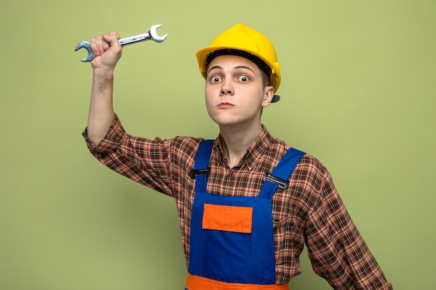 Frowning young male builder wearing uniform holding open-end wrench 