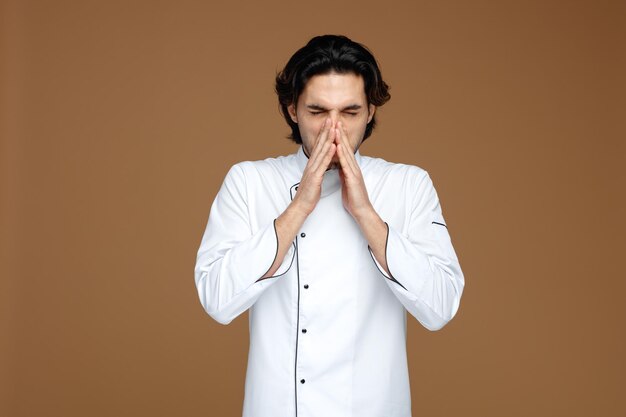 frowning young male chef wearing uniform keeping hands on nose sneezing with closed eyes isolated on brown background