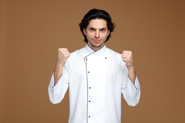 хмурый молодой шеф-повар в униформе смотрит в камеру, показывая кулаки в камеру на коричневом фоне