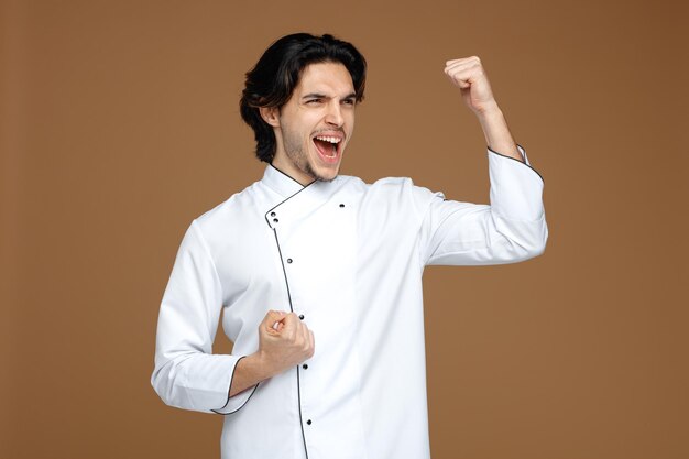 frowning young male chef wearing uniform looking at side showing yes gesture isolated on brown background