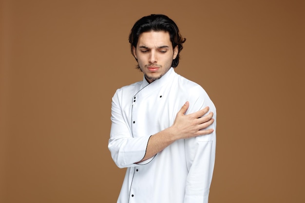 Accigliato giovane chef maschio che indossa l'uniforme in piedi nella vista di profilo tenendo la mano sul braccio guardando in basso isolato su sfondo marrone con spazio per la copia