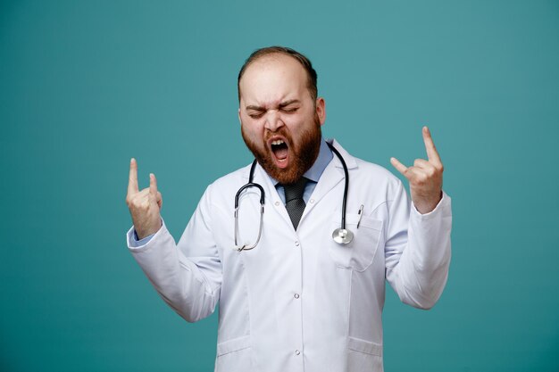 Frowning young male doctor wearing medical coat and stethoscope around his neck showing rock sign screaming with closed eyes isolated on blue background