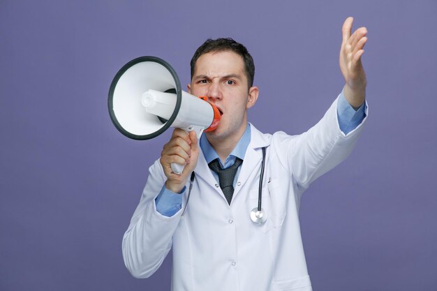 Frowning young male doctor wearing medical robe and stethoscope around neck looking at camera keeping hand in air talking into laudspeaker isolated on purple background