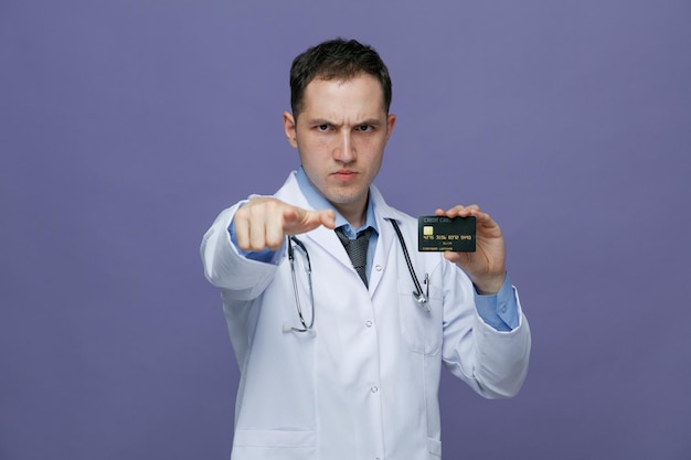 紫色の背景に分離されたカメラを指しているクレジットカードを示すカメラを見て首の周りに医療ローブと聴診器を身に着けている若い男性医師を慌てて