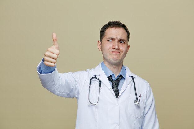 Frowning young male doctor wearing medical robe and stethoscope around neck looking at side showing thumb up isolated on olive green background