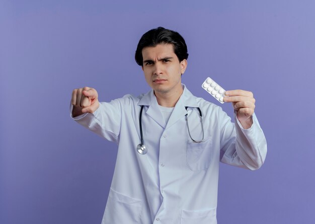 Frowning young male doctor wearing medical robe and stethoscope showing pack of tablets looking and pointing  isolated on purple wall with copy space