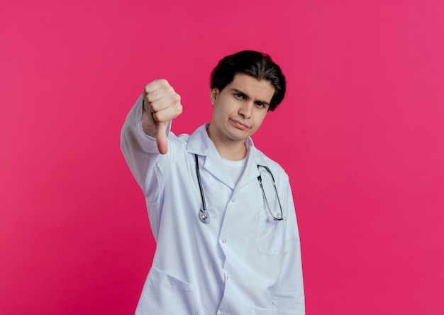 Хмурый молодой мужчина-врач в медицинском халате и стетоскопе показывает большой палец вниз на розовой стене с копией пространства