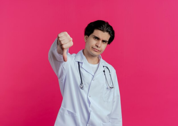 Frowning young male doctor wearing medical robe and stethoscope  showing thumb down isolated on pink wall with copy space