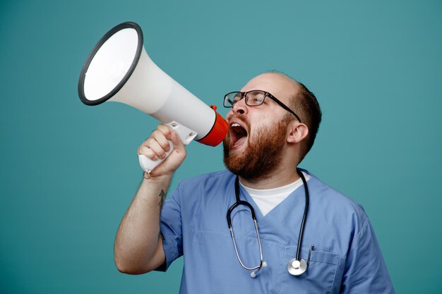 frowning young male nurse wearing glasses nurse scrub and stethoscope around his neck looking at side talking into speaker isolated on blue background