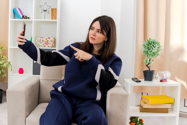 Frowning young pretty caucasian woman sitting on armchair in designed living room pointing at phone and taking selfie