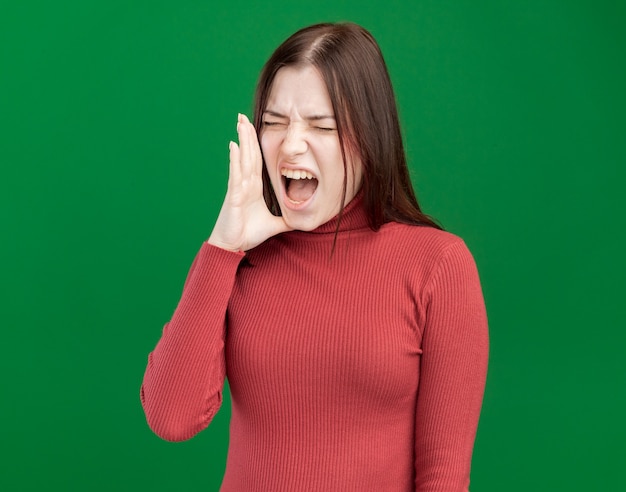 Frowning young pretty woman keeping hand near mouth calling out loud to someone with closed eyes isolated on green wall