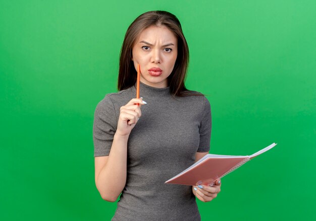 Frowning young pretty woman touching face with pen and holding open note pad isolated on green background with copy space