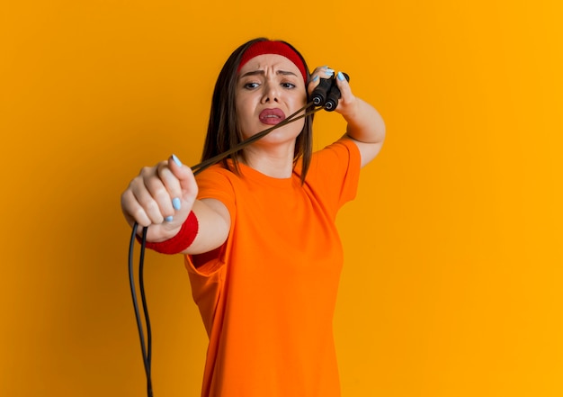 Нахмурившаяся молодая спортивная женщина в головной повязке и браслетах тренируется со скакалкой, глядя на нее, изолированную на оранжевой стене с копией пространства
