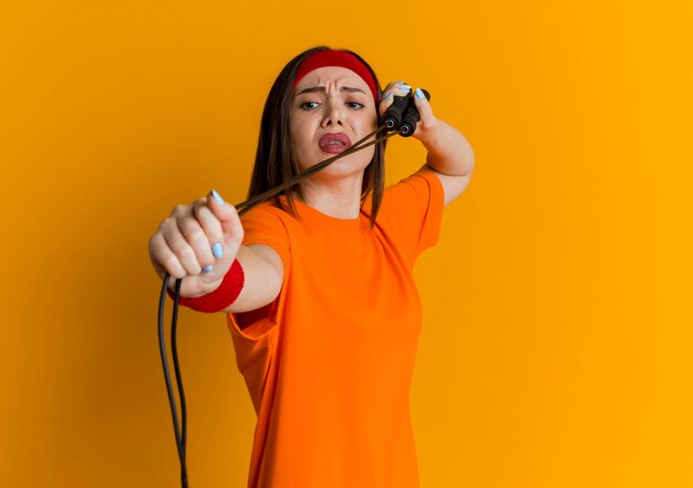 Frowning young sporty woman wearing headband and wristbands exercising with jump rope looking at it isolated on orange wall with copy space