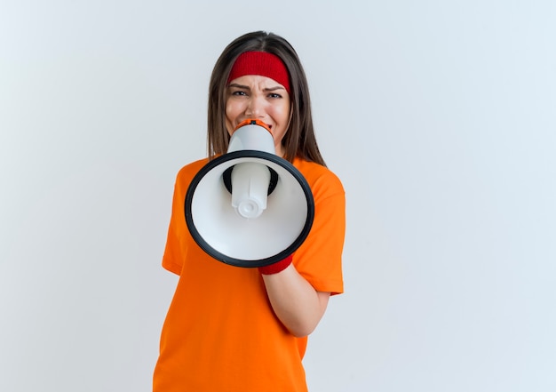 Frowning young sporty woman wearing headband and wristbands looking talking by speaker isolated