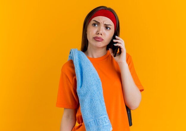 Frowning young sporty woman wearing headband and wristbands with jump rope and towel on shoulders talking on phone looking at side 
