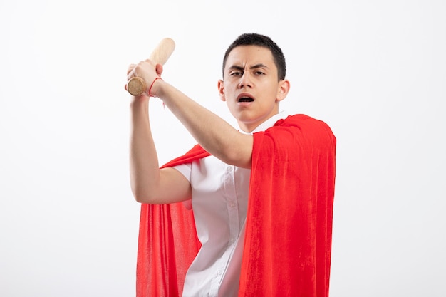Frowning young superhero boy in red cape raising baseball bat up looking at camera getting ready to hit isolated on white background with copy space