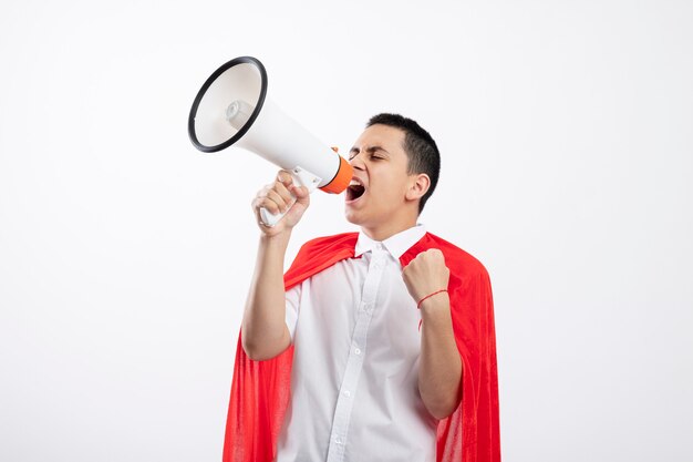 Frowning young superhero boy in red cape shouting in loud speaker with closed eyes clenching fist isolated on white background with copy space