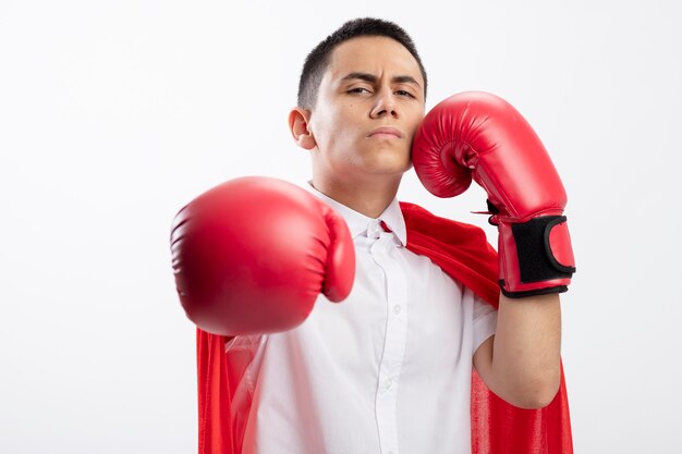 Frowning young superhero boy in red cape wearing box gloves looking at camera stretching out hand towards camera touching face with another one isolated on white background with copy space