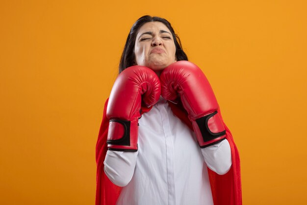 Frowning young superwoman wearing box gloves keeping hands under chin with closed eyes isolated on orange wall