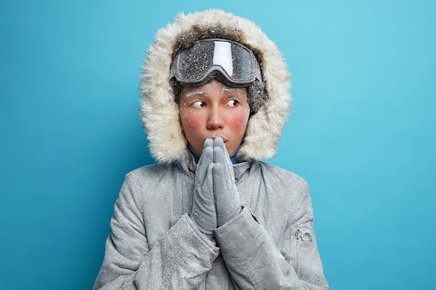 Frozen young ethnic woman tries to warm after spending long time at cold day keeps hands pressed together blows warm air wears grey jacket with hood has cold face covered with white hoarfrost