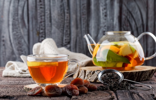 Fruit infused water in teapot with tea, dried apricots, wood, kitchen towel, container side view on stone tile and wooden surface