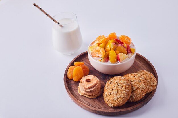 Fruit salad in cream with oatmeal cookies in a wooden plate with a glass of milk aside, top view.