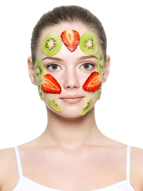 白で隔離される美しい女性の若い顔にイチゴとキウイのフルーツイチゴマスク