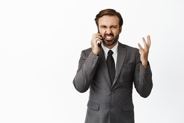 Разочарованный и сердитый предприниматель спорит во время разговора с интенсивным телефонным звонком, стоящим на белом фоне