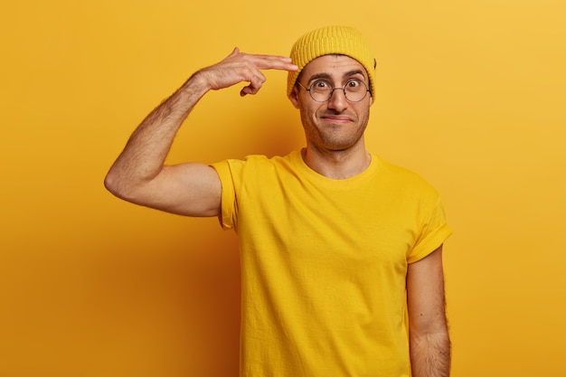 Frustrated adult male shows gun pistole, keeps fingers near temple, frowns face, distressed with much work to do, wears yellow hat and t-shirt