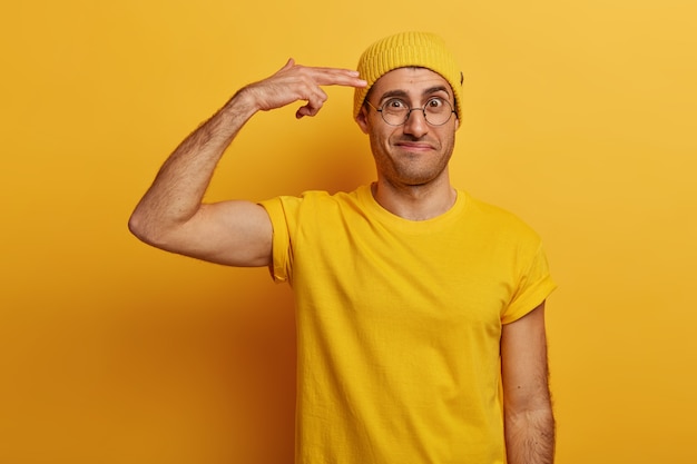 Разочарованный взрослый мужчина показывает пистолетный пистолет, держит пальцы у виска, хмурится, огорченный большой работой, носит желтую шляпу и футболку