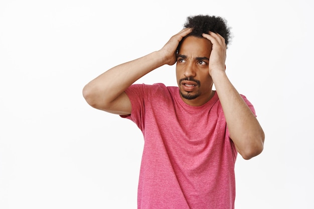 Frustrated african american man holding hands on head, looking aside at banner with troubled, concerned and shocked look, overwhelmed by smth alarming, white background