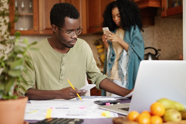 Frustrated African husband in glasses holding cell phone and pencil, managing family expenses and paying domestic bills online