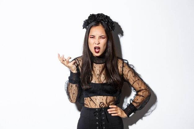 Frustrated and angry young asian woman in witch dress arguing with someone, looking pissed-off and bothered, shaking hand and looking scary at camera, standing over white background.