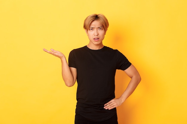 Frustrated and annoyed asian guy with blond hair, raising hand confused, yellow wall