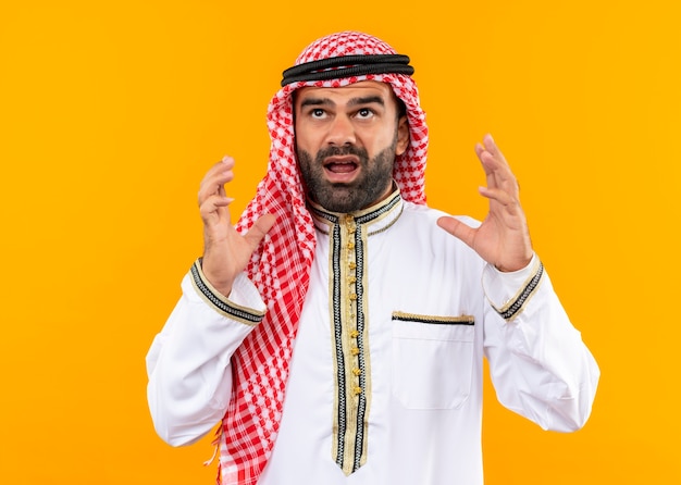 Frustrated arabic businessman in traditional wear shouting with raised hands standing over orange wall