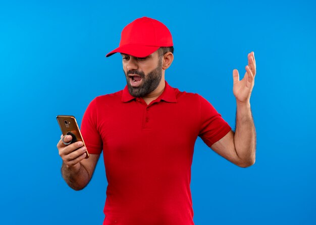 Разочарованный бородатый курьер в красной униформе и кепке с поднимающей руку со смартфоном недоволен, стоя у синей стены