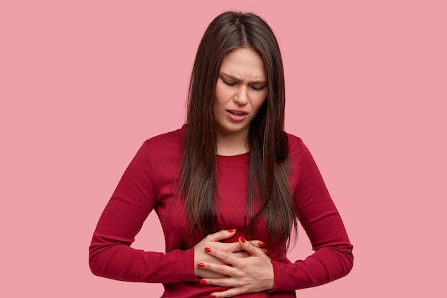 Frustrated brunette woman with displeased expression, keeps hands on belly, feels pain as has menstruation, wears red sweater