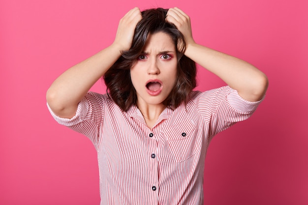 Frustrated confused Caucasian woman keeping hands in her hair, posing with widely opened mouth, wearing striped shirt, being in bad mood, looks shocked.