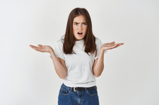 Frustrated and confused woman asking why, have questions and cant understand something, shrugging shoulders and staring at front, white wall