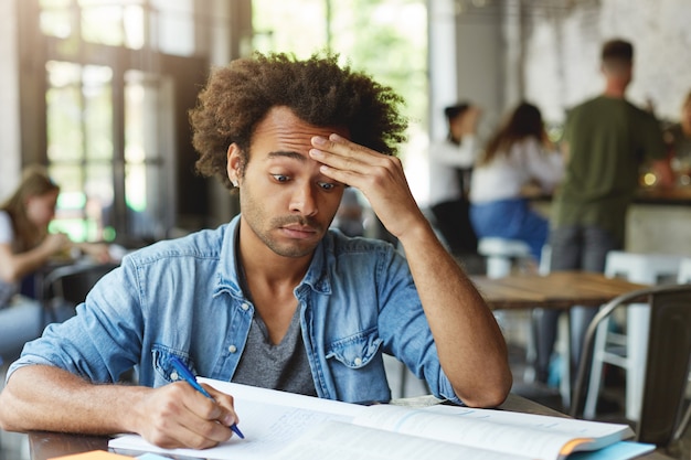 Frustrated confused young college student with Afro hairstyle rubbing forehead, trying hard to understand complicated mathematical problem while doing homework at cafe, using pen for making notes