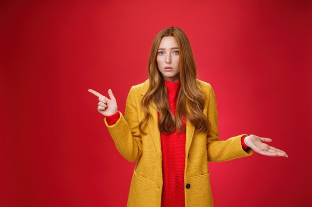 Frustrated and confused young cute female with ginger hair in yellow coat shrugging as pointing questioned left waiting explanation as asking question clueless and puzzled over red background.