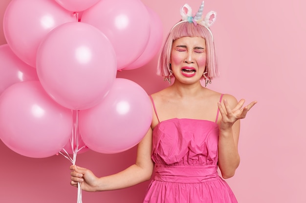 Free photo frustrated crying asian woman with pink bob hair raises hand has mournful face expression upset because of holiday celebration alone