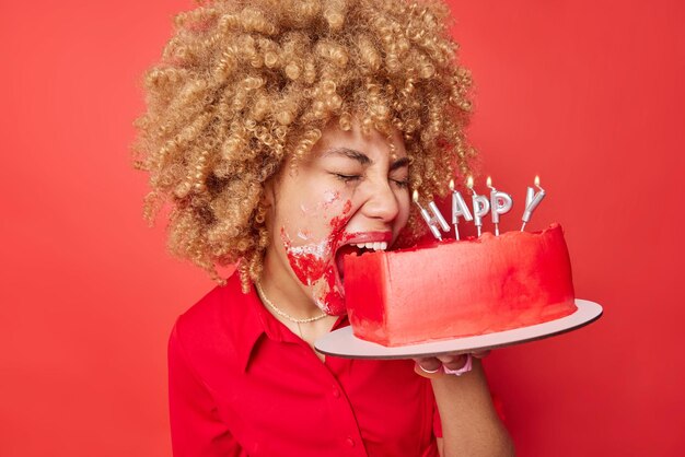 크림으로 얼룩진 케이크를 물고 있는 좌절한 곱슬머리 여성은 발렌타인 데이를 축하하기만 하면 생생한 붉은 배경에 대해 우울한 기분이 드는 포즈를 취하고 있습니다. 버릇없는 휴가와 외로운 축하 개념
