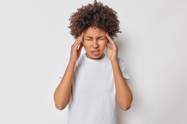 Frustrated curly haired woman keeps hands on temples suffers from migraine has headache dressed in casual t shirt isolated over white background feels discomfort grimaces from pain or disappointment