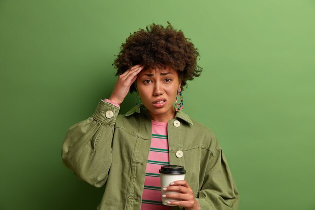 Frustrated curly haired woman suffers from headache, touches temple, drinks refreshing beverage after sleepless night, holds disposable cup of coffee, dressed in stylish outfit, isolated on green wall