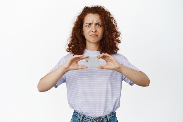 Frustrated curly woman frowning, grimacing upset, showing credit discount card, standing in summer t-shirt on white