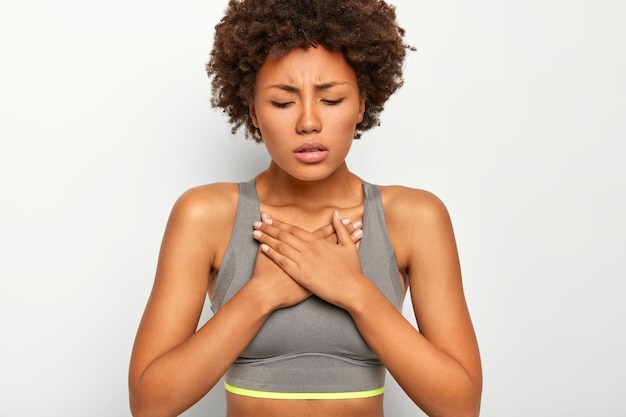 Frustrated dark skinned Afro American woman suffers from acute pain in chest, wears grey sport bra, isolated on white background