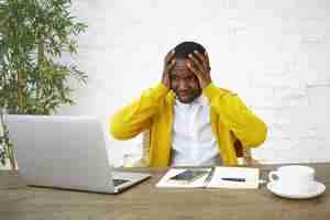 Free photo frustrated dark skinned businessman sitting at workplace with hands on head, feeling stressed, looking at laptop screen in panic, unable to keep company afloat, having not enough money to run business