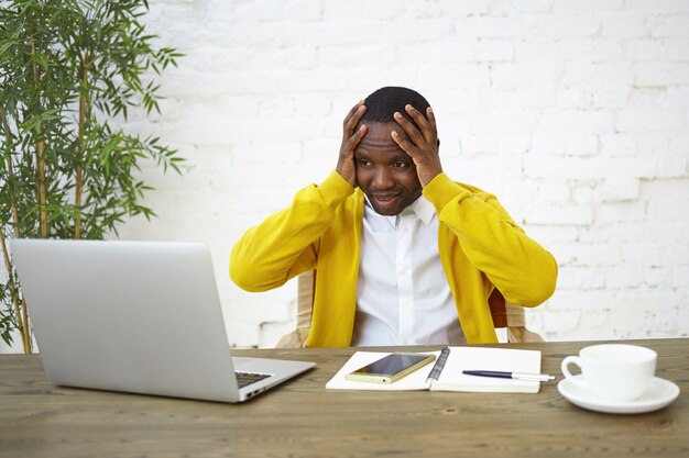 Frustrated dark skinned businessman sitting at workplace with hands on head, feeling stressed, looking at laptop screen in panic, unable to keep company afloat, having not enough money to run business